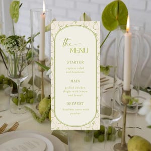 Puede incluir: Una mesa elegante con una tarjeta de menú, velas y arreglos florales. La tarjeta de menú muestra "The MENU" y una lista de platos. Velas altas en portavelas de vidrio y follaje verde contribuyen al ambiente.