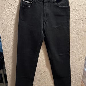 Könnte beinhalten: Eine schwarze Jeans aus Denim hängt an einer strukturierten Wand. Die Jeans hat ein klassisches Fünf-Taschen-Design, einen Knopf- und Reißverschluss und ein kleines Logo-Detail auf der Vordertasche.