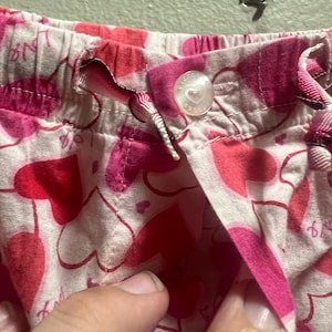 以下が含まれることがあります： ピンクと赤のハート柄のパジャマショートパンツのクローズアップ。 ショーツは、ドローストリングウエストバンドとハートのデザインのボタン留めです。 生地は、繰り返しのハート柄の薄いピンク色です。
