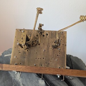 May include: A vintage clock movement with brass gears and levers mounted on a rectangular brass plate. The plate is attached to a wooden base. The clock mechanism has a complex design with intricate details.