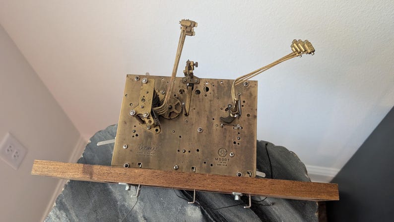 May include: A vintage clock movement with brass gears and levers mounted on a rectangular brass plate. The plate is attached to a wooden base. The clock mechanism has a complex design with intricate details.