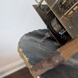 May include: Close-up of a vintage clock mechanism. The brass-colored gears and plates are partially visible, along with a wooden pendulum. The clock rests on a dark gray, textured surface with gold accents.