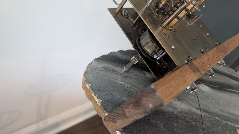 May include: Close-up of a vintage clock mechanism. The brass-colored gears and plates are partially visible, along with a wooden pendulum. The clock rests on a dark gray, textured surface with gold accents.