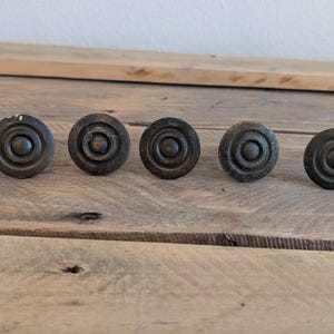 Könnte beinhalten: Sechs antike Schrankknöpfe aus Bronze mit kreisförmigem Design sind auf einer verwitterten Holzoberfläche angeordnet. Die Knöpfe haben ein dunkles, gealtertes Aussehen, was darauf hindeutet, dass sie Vintage oder recycelt sind. Die Knöpfe sind ein dekoratives Hardware-Element.