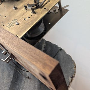 May include: Close-up of a vintage clock mechanism. The image shows a wooden frame with metal hooks and wires, attached to a brass-colored plate with gears and other components. The clock's base is a dark gray color.