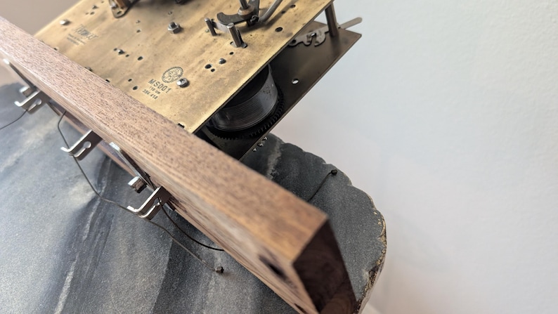 May include: Close-up of a vintage clock mechanism. The image shows a wooden frame with metal hooks and wires, attached to a brass-colored plate with gears and other components. The clock's base is a dark gray color.