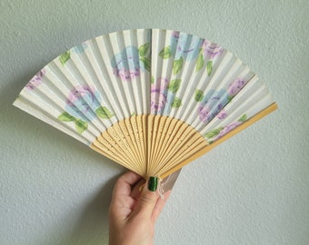 Puede incluir: Un abanico plegable blanco con un estampado floral en tonos azules, morados y verdes. El mango de madera del abanico es de color marrón claro. El abanico se sostiene abierto, mostrando su diseño completo.