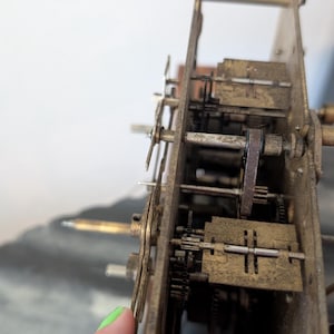 May include: Close-up of a vintage clock mechanism, showcasing intricate gears, levers, and springs. The aged brass components are visible, with a focus on the mechanical details. The clock's internal structure is partially exposed, revealing its complex design.