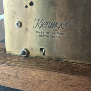 May include: Close-up of a brass clock mechanism with the name "Kieninger" and "Made in Germany" visible. The serial number 9823818639 is also present. The mechanism is mounted on a wooden base.