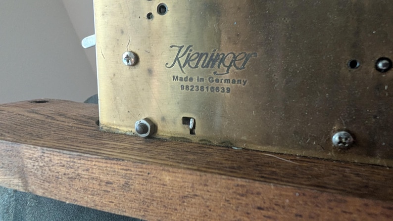 May include: Close-up of a brass clock mechanism with the name "Kieninger" and "Made in Germany" visible. The serial number 9823818639 is also present. The mechanism is mounted on a wooden base.