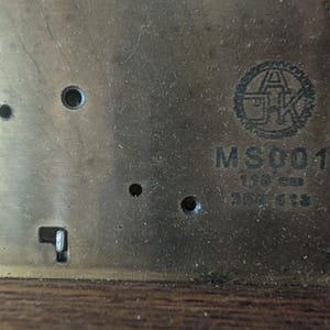 May include: Close-up of a brass metal plate with the text "MS001" and other markings. The plate has several screw holes and a circular logo. The text "150 cm" is also visible.