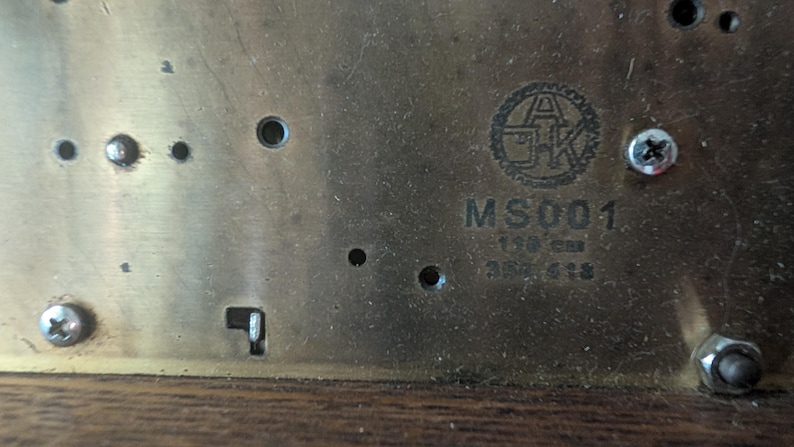 May include: Close-up of a brass metal plate with the text "MS001" and other markings. The plate has several screw holes and a circular logo. The text "150 cm" is also visible.