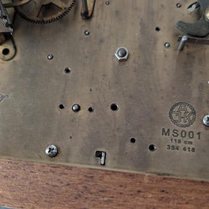 May include: Close-up of a clock mechanism with brass components. The text "Kieninger Made in Germany" is visible, along with the model number "MS001" and dimensions of 118 cm. The clock's gears and screws are also visible.