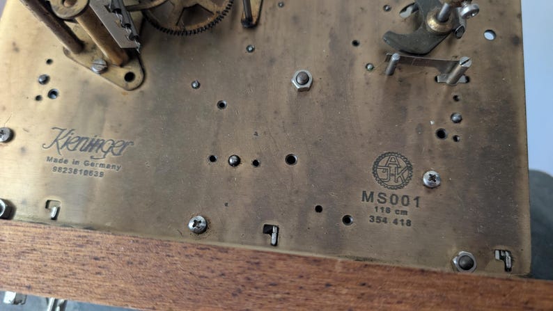 May include: Close-up of a clock mechanism with brass components. The text "Kieninger Made in Germany" is visible, along with the model number "MS001" and dimensions of 118 cm. The clock's gears and screws are also visible.