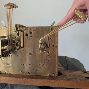 May include: A vintage clock movement with brass gears and components. The clock mechanism is mounted on a wooden base. The image shows the intricate details of the clock's internal workings, including gears, levers, and pendulums. The clock is marked "Kieninger" and "Made in Germany".