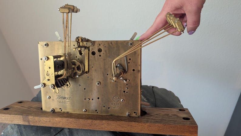 May include: A vintage clock movement with brass gears and components. The clock mechanism is mounted on a wooden base. The image shows the intricate details of the clock's internal workings, including gears, levers, and pendulums. The clock is marked "Kieninger" and "Made in Germany".