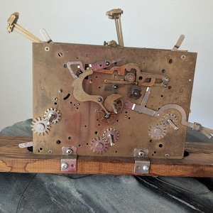 May include: A vintage clock mechanism with gears, levers, and a brass plate. The clock's internal components are mounted on a wooden base. The mechanism is a complex arrangement of metal parts, showcasing the inner workings of a clock.