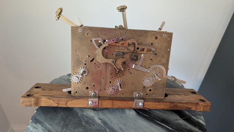 May include: A vintage clock mechanism with gears, levers, and a brass plate. The clock's internal components are mounted on a wooden base. The mechanism is a complex arrangement of metal parts, showcasing the inner workings of a clock.