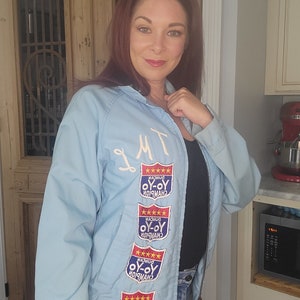 May include: Light blue jacket with embroidered patches that read "Duncan Yo-Yo Champion." The jacket has a zipper closure and white embroidered initials. The patches are red, white, and blue with gold stars.