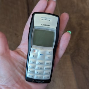 Op de afbeelding: Een zwart en zilver Nokia mobiele telefoon met een toetsenbord en scherm. De telefoon wordt in een hand gehouden. De telefoon heeft de merknaam "Nokia" bovenaan afgedrukt.