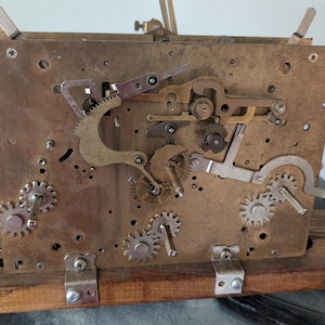 May include: Close-up of a vintage clock mechanism. The brass-colored plate features various gears, levers, and screws. The mechanism is mounted on a wooden base. The clock's internal components are visible, showcasing its intricate design.