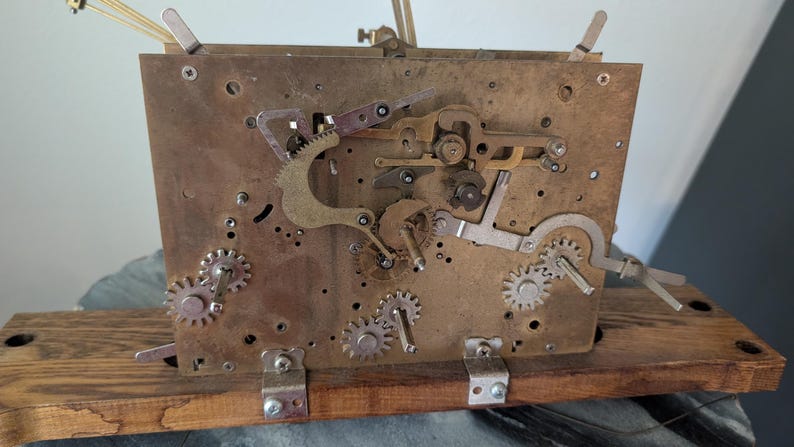 May include: Close-up of a vintage clock mechanism. The brass-colored plate features various gears, levers, and screws. The mechanism is mounted on a wooden base. The clock's internal components are visible, showcasing its intricate design.
