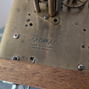 May include: Close-up of a brass clock mechanism with the name "Kieninger" and "Made in Germany" visible. The mechanism has various gears, screws, and other components. A wooden support is visible at the bottom of the image.