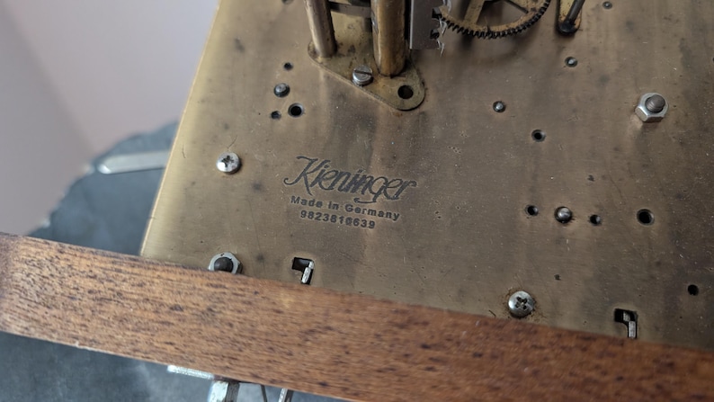 May include: Close-up of a brass clock mechanism with the name "Kieninger" and "Made in Germany" visible. The mechanism has various gears, screws, and other components. A wooden support is visible at the bottom of the image.
