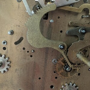 May include: Close-up of a clock mechanism with various gears, levers, and metal components. The gears are silver and gold, and the background is a brass-colored plate. The image shows the intricate details of the clock's internal workings.