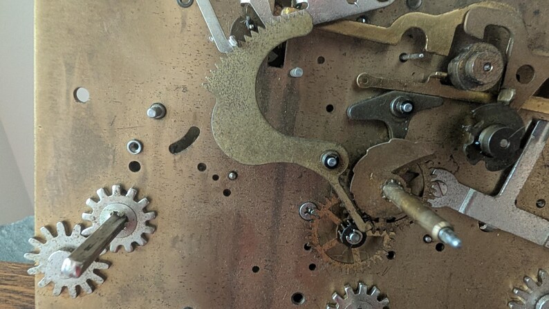May include: Close-up of a clock mechanism with various gears, levers, and metal components. The gears are silver and gold, and the background is a brass-colored plate. The image shows the intricate details of the clock's internal workings.