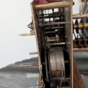 May include: Close-up of a vintage clock mechanism. The intricate internal workings are visible, featuring gears, springs, and other components. The metal parts have a weathered, aged appearance, suggesting the clock's age. The wooden base is also visible.