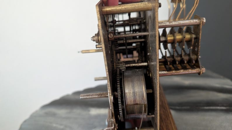 May include: Close-up of a vintage clock mechanism. The intricate internal workings are visible, featuring gears, springs, and other components. The metal parts have a weathered, aged appearance, suggesting the clock's age. The wooden base is also visible.