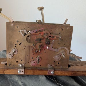 May include: A vintage clock movement with brass gears, levers, and a wooden base. The mechanism features multiple gears, levers, and a balance wheel. The clock is mounted on a rectangular wooden base, and the overall color scheme is a mix of brass, copper, and wood.