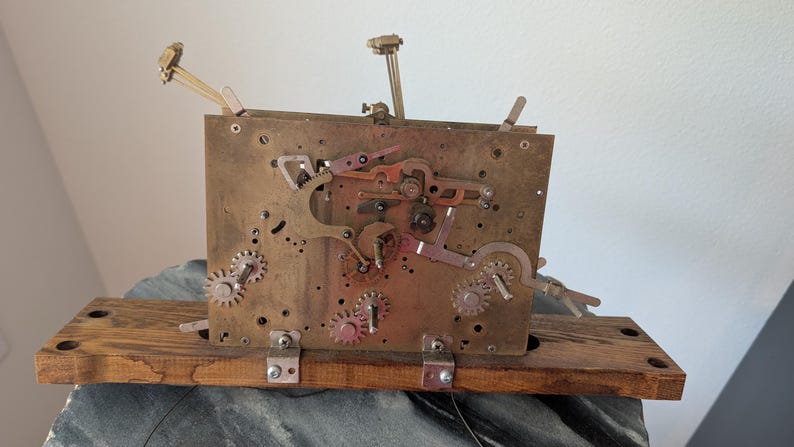 May include: A vintage clock movement with brass gears, levers, and a wooden base. The mechanism features multiple gears, levers, and a balance wheel. The clock is mounted on a rectangular wooden base, and the overall color scheme is a mix of brass, copper, and wood.