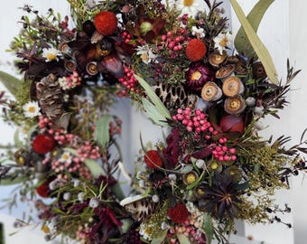 May include: A circular wreath composed of dried botanical elements including dark purple protea, bright red berries, small white flowers, eucalyptus leaves, brown seed pods, and various textured twigs arranged in a dense, multi-layered display.