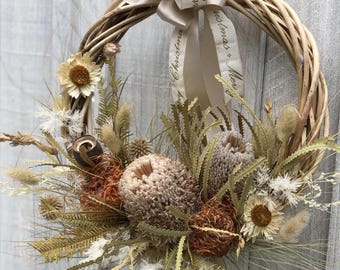 May include: A natural wreath made of woven branches, adorned with dried flowers and foliage in shades of brown, beige, and green. A cream-colored ribbon with the words "Merry Christmas" is tied at the top. This decorative wreath is perfect for seasonal home decor.
