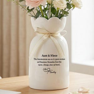 May include: A white ceramic vase with a scalloped top, tied with a cream ribbon, holds a bouquet of pink and white flowers and greenery. The vase has text that reads "Aunt & Niece".