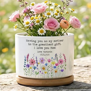 Puede incluir: Una maceta de cerámica blanca con base de bambú, llena de rosas rosas, margaritas y otras flores coloridas. La maceta tiene un diseño floral y el texto "Having you as my mother is the greatest gift. I love you Mom".