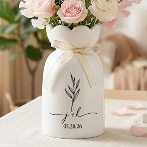 Pode incluir: Um vaso de cerâmica branco com a parte superior ondulada, cheio de flores rosa e brancas. O vaso tem um design floral preto e a inscrição "j + h 05.20.26" com uma fita creme em volta do pescoço. O vaso está numa mesa.