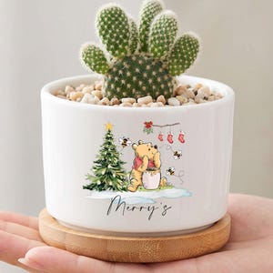 May include: A white ceramic cylindrical planter with a bamboo saucer base containing a small green cactus and light-colored pebbles. The planter features a graphic of a bear with a honey pot, a Christmas tree, and the text Merry's in black script.
