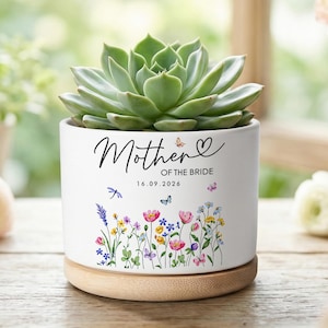 Könnte beinhalten: Ein weißer Keramik-Pflanztopf mit einer Sukkulente, mit der Aufschrift "Mother of the Bride" und dem Datum 16.09.2026. Der Topf ist mit floralen und Schmetterlingsillustrationen verziert und steht auf einem Holzsockel.