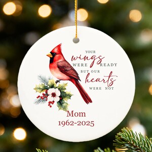 Può includere: Ornamento natalizio rotondo in ceramica bianca con un'illustrazione di un cardinale rosso, agrifoglio e testo. Il testo recita: "Le tue ali erano pronte ma i nostri cuori no". Sotto il testo c'è "Mamma 1962-2025".