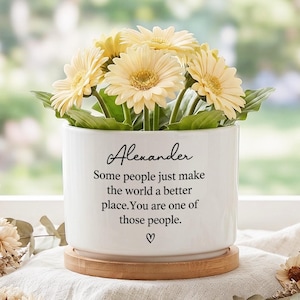 May include: A white ceramic plant pot with a wooden base, filled with yellow gerbera daisies. The pot has the name "Alexander" and a message printed in black. A heart symbol is at the bottom.