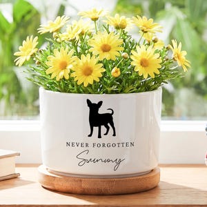 Op de afbeelding: Witte keramische bloempot met een zwart silhouet van een hond, gevuld met gele bloemen. De pot heeft de tekst "NEVER FORGOTTEN" en de naam "Sunny". De pot staat op een houten schotel.