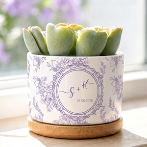 May include: A white ceramic planter with a light purple floral pattern and a central circular emblem featuring the letters S + K and the date 07.06.2026. The planter sits on a round bamboo saucer and contains a green succulent plant.