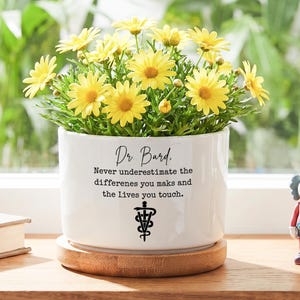 May include: A white ceramic plant pot filled with yellow flowers, on a wooden saucer. The pot features the text "Dr. Bard. Never underestimate the differences you make and the lives you touch." and a medical symbol.