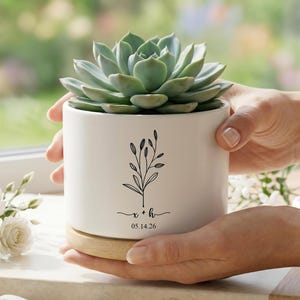 May include: A white ceramic planter with a succulent plant. The planter has a wooden base and a black floral design with the initials "x + h" and the date "05.14.26". The succulent has green leaves.