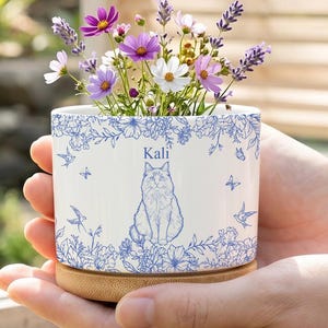 Puede incluir: Una maceta de cerámica blanca con un diseño floral azul y una ilustración de gato, llena de flores coloridas. La maceta tiene el nombre "Kali" impreso y se asienta sobre una base de madera.