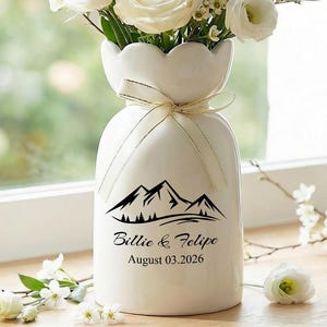May include: A white ceramic vase with a scalloped top, tied with a ribbon, holds a bouquet of white roses and greenery. The vase features a mountain graphic and the names "Billie & Felipe" with the date "August 03,2026".