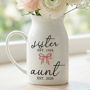 May include: A white ceramic vase with a handle, filled with pink and white flowers. The vase has the words "sister" and "aunt" in script, with dates and a pink bow graphic. The vase is on a wooden surface.